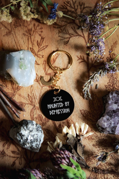 Ein Schlüsselanhänger in Schwarz und Gold mit goldenem Mond-Charm. Auf der schwarzen Scheibe steht in weiß: Haunted by depression