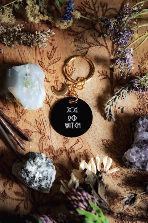 Ein Schlüsselanhänger in Schwarz und Gold mit goldenem Mond-Charm. Auf der schwarzen Scheibe steht in weiß: OCD Witch
