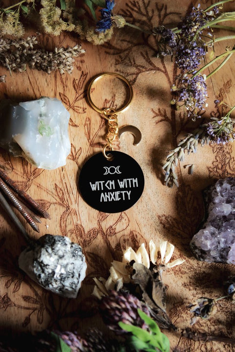 Ein Schlüsselanhänger in Schwarz und Gold mit goldenem Mond-Charm. Auf der schwarzen Scheibe steht in weiß: Witch with Anxiety