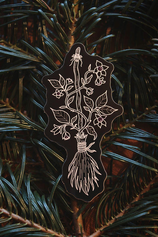 Auf Tannennadeln gebettet liegt ein schwarzer Aufkleber mit weißer Zeichnung eines Hexenbesens um den Tollkirsche geschlungen ist.