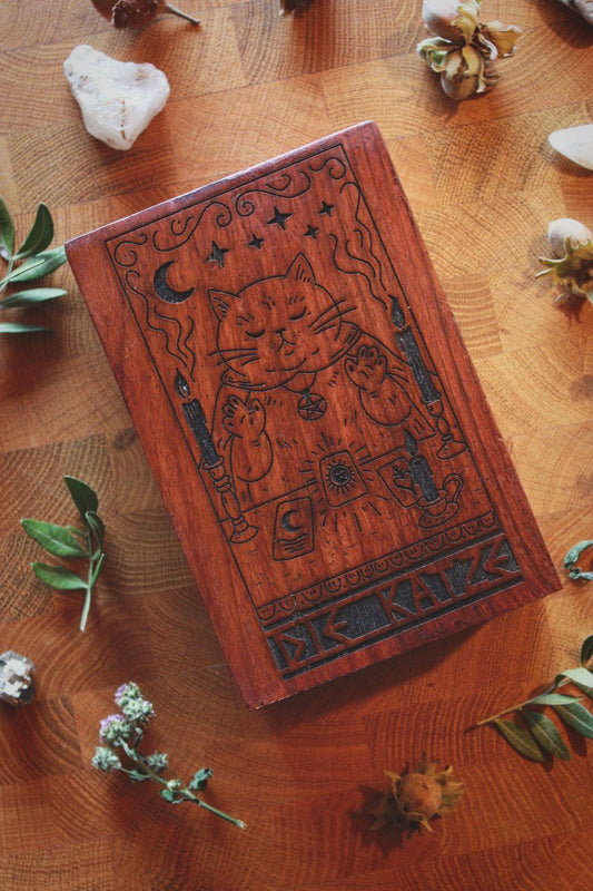 Holzkiste für Tarotkarten mit eingebranntem Motiv: Eine ruhende, mystisch wirkende Katze sitzt zwischen zwei brennenden Kerzen und legt Tarotkarten. Über ihr schweben Sterne und ein Mond, darunter steht ‚DIE KATZE‘. Die Szene ist von Blättern, Steinen und Naturmaterialien umgeben.

