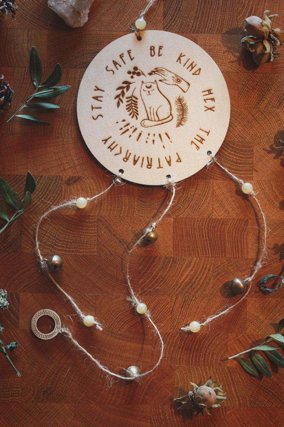 Rundes Holzschild mit Gravur: Eine Katze mit einer schützenden Hand über ihr. Darunter steht der Spruch: „STAY SAFE BE KIND HEX THE PATRIARCHY“. Das Schild ist mit Perlen aus Aragonit, Glöckchen und einer Schlange verziert.
