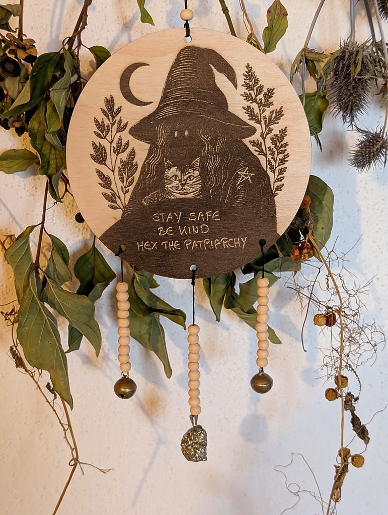 Rundes Holzschild mit Gravur einer Hexe mit spitzem Hut und Katze im Arm, umgeben von Blättern und einem Halbmond. Darunter steht der Spruch: „STAY SAFE BE KIND HEX THE PATRIARCHY“. Das Schild ist mit Perlen, zwei Glöckchen und Pyrit verziert und hängt vor grünem Blattwerk.