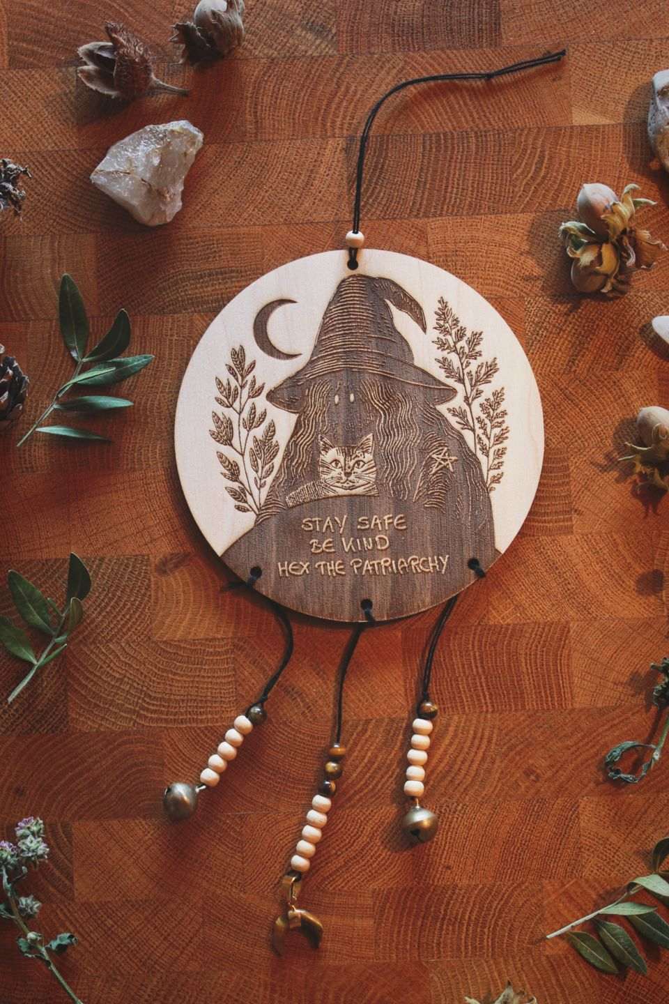 Rundes Holzschild mit Gravur einer Hexe mit spitzem Hut und Katze im Arm, umgeben von Blättern und einem Halbmond. Darunter steht der Spruch: „STAY SAFE BE KIND HEX THE PATRIARCHY“. Das Schild ist mit Perlen, zwei Glöckchen und einem halbmondförmigen Tigerauge verziert.
