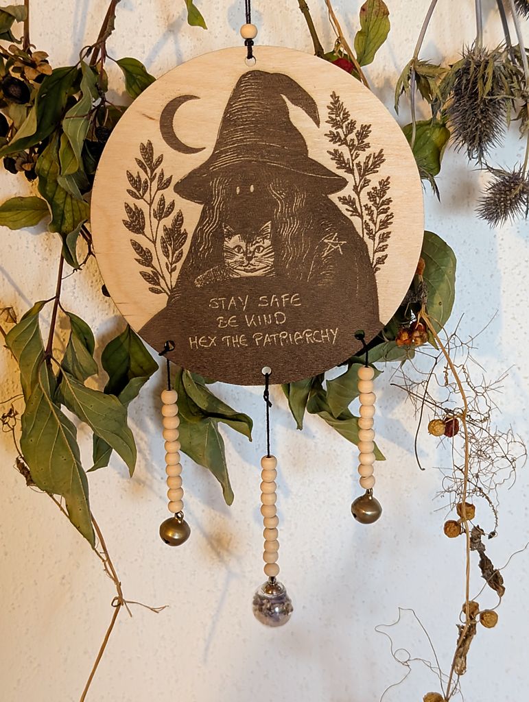 Rundes Holzschild mit Gravur einer Hexe mit spitzem Hut und Katze im Arm, umgeben von Blättern und einem Halbmond. Darunter steht der Spruch: „STAY SAFE BE KIND HEX THE PATRIARCHY“. Das Schild ist mit Perlen, zwei Glöckchen und einer Kugel gefüllt mit Blauraute und Rosenquarz verziert und hängt vor grünem Blattwerk.