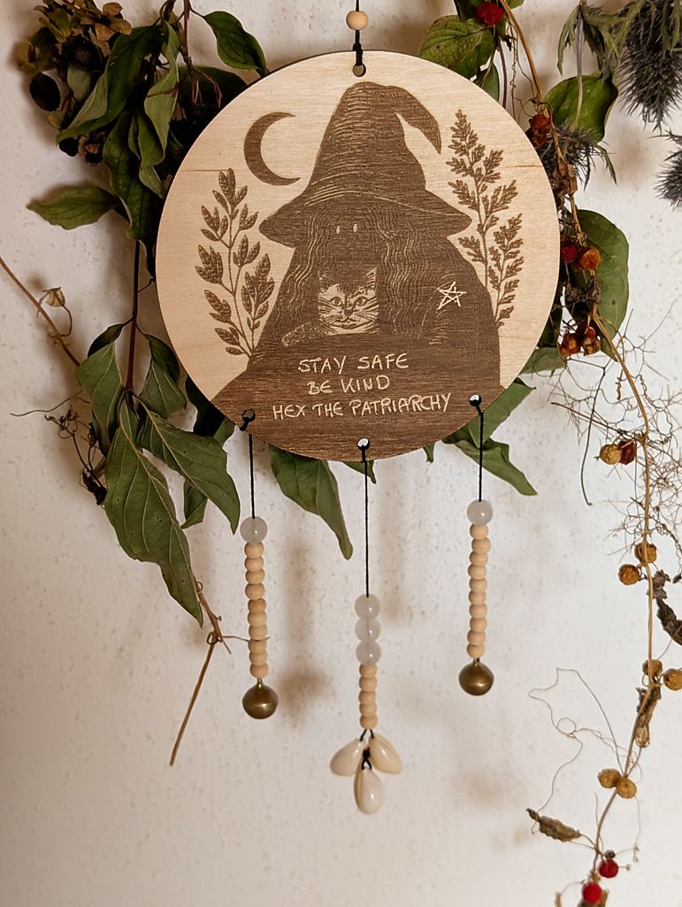 Rundes Holzschild mit Gravur einer Hexe mit spitzem Hut und Katze im Arm, umgeben von Blättern und einem Halbmond. Darunter steht der Spruch: „STAY SAFE BE KIND HEX THE PATRIARCHY“. Das Schild ist mit Perlen, zwei Glöckchen und drei Muscheln verziert und hängt vor grünem Blattwerk.