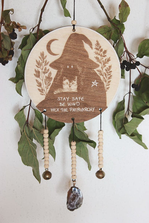 Rundes Holzschild mit Gravur einer Hexe mit spitzem Hut und Katze im Arm, umgeben von Blättern und einem Halbmond. Darunter steht der Spruch: „STAY SAFE BE KIND HEX THE PATRIARCHY“. Das Schild ist mit Perlen, zwei Glöckchen und einer Achat-Geode verziert und hängt vor grünem Blattwerk.