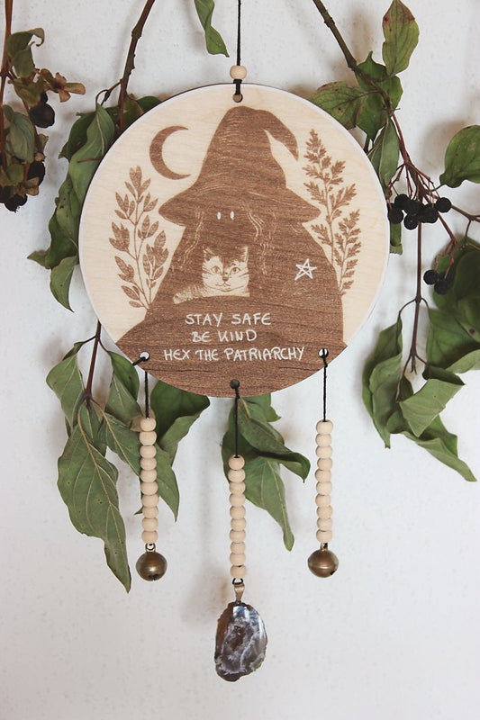 Rundes Holzschild mit Gravur einer Hexe mit spitzem Hut und Katze im Arm, umgeben von Blättern und einem Halbmond. Darunter steht der Spruch: „STAY SAFE BE KIND HEX THE PATRIARCHY“. Das Schild ist mit Perlen, zwei Glöckchen und einer Achat-Geode verziert und hängt vor grünem Blattwerk.
