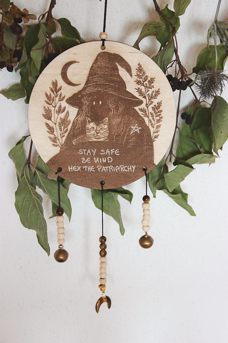 Rundes Holzschild mit Gravur einer Hexe mit spitzem Hut und Katze im Arm, umgeben von Blättern und einem Halbmond. Darunter steht der Spruch: „STAY SAFE BE KIND HEX THE PATRIARCHY“. Das Schild ist mit Perlen, zwei Glöckchen und einem halbmondförmigen Tigerauge verziert und hängt vor grünem Blattwerk.