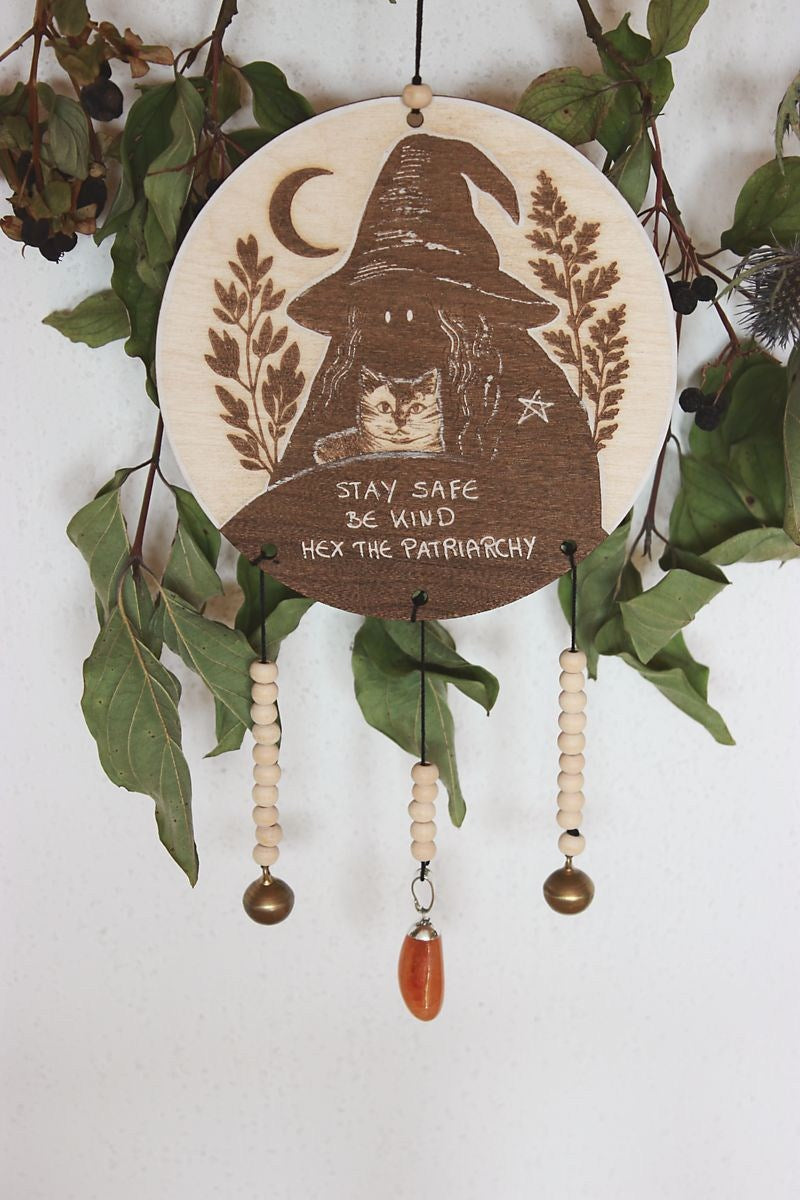Rundes Holzschild mit Gravur einer Hexe mit spitzem Hut und Katze im Arm, umgeben von Blättern und einem Halbmond. Darunter steht der Spruch: „STAY SAFE BE KIND HEX THE PATRIARCHY“. Das Schild ist mit Perlen, zwei Glöckchen und einem Karneol verziert und hängt vor grünem Blattwerk.
