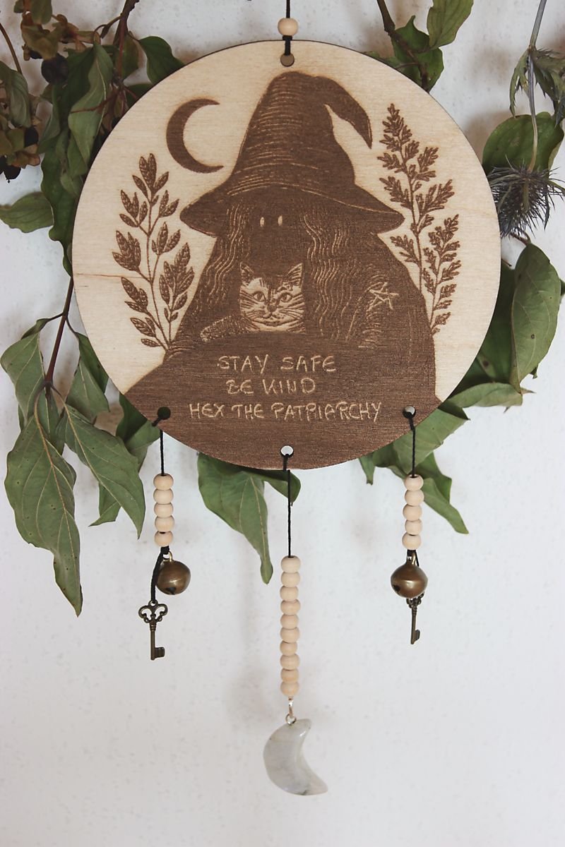 Rundes Holzschild mit Gravur einer Hexe mit spitzem Hut und Katze im Arm, umgeben von Blättern und einem Halbmond. Darunter steht der Spruch: „STAY SAFE BE KIND HEX THE PATRIARCHY“. Das Schild ist mit Perlen, zwei Schlüsseln, zwei Glöckchen und einem halbmondförmigen Stein verziert und hängt vor grünem Blattwerk.