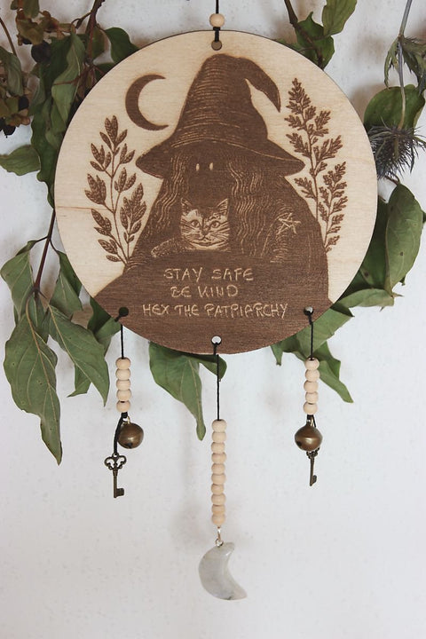 Rundes Holzschild mit Gravur einer Hexe mit spitzem Hut und Katze im Arm, umgeben von Blättern und einem Halbmond. Darunter steht der Spruch: „STAY SAFE BE KIND HEX THE PATRIARCHY“. Das Schild ist mit Perlen, zwei Schlüsseln, zwei Glöckchen und einem halbmondförmigen Stein verziert und hängt vor grünem Blattwerk.