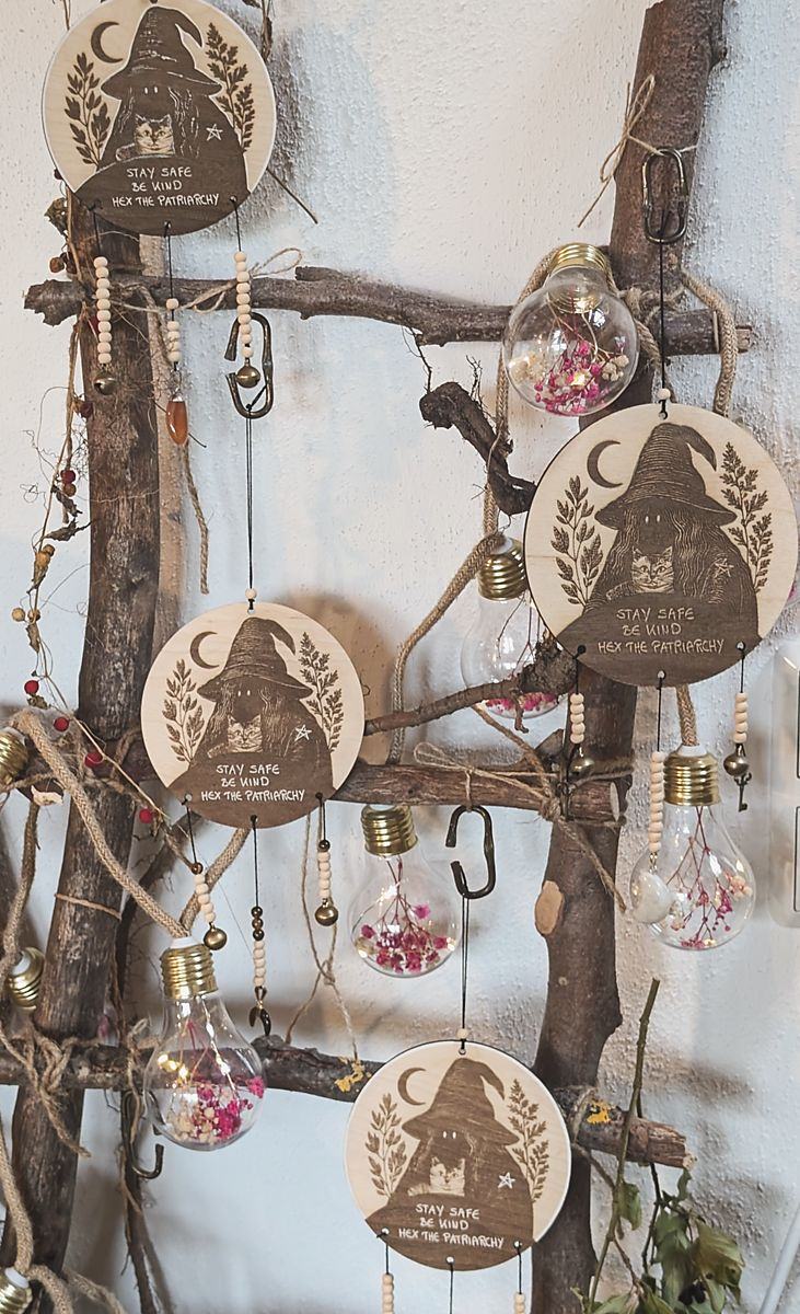 Dekoratives Arrangement mit Holzleiter, an der runde Holzschilder und Glühbirnen mit rosa-weißen Trockenblumen hängen. Die Schilder zeigen eine Figur mit Hut, botanische Elemente und den Text: „STAY SAFE BE KIND HEX THE PATRIARCHY“. Natürliche Materialien und liebevolle Details verleihen dem Ensemble einen rustikalen, achtsamen Charakter.