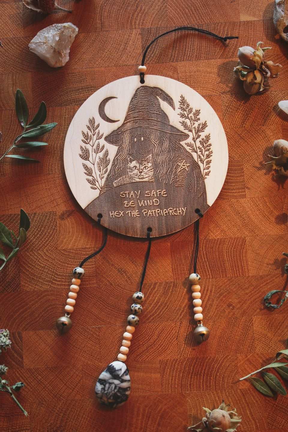 Rundes Holzschild mit Gravur einer Hexe mit spitzem Hut und Katze im Arm, umgeben von Blättern und einem Halbmond. Darunter steht der Spruch: „STAY SAFE BE KIND HEX THE PATRIARCHY“. Das Schild ist mit Perlen, zwei Glöckchen und Eisblumen-Magnesit sowie Jaspis verziert.