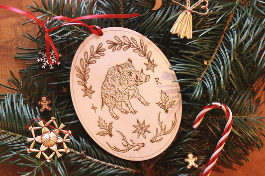 Gebettet auf Tannenzweigen und weihnachtliche Dekoration liegt in der Mitte ein ovales Ornament aus Holz. Diese Dekoration zu Jul (Wintersonnenwende) zeigt ein Wildschwein. Eingerahmt wird es von typischen Jul-Motiven wie Mistelzweigen, Stechpalmen, Strohsternen, Geweihen und passenden Runen.