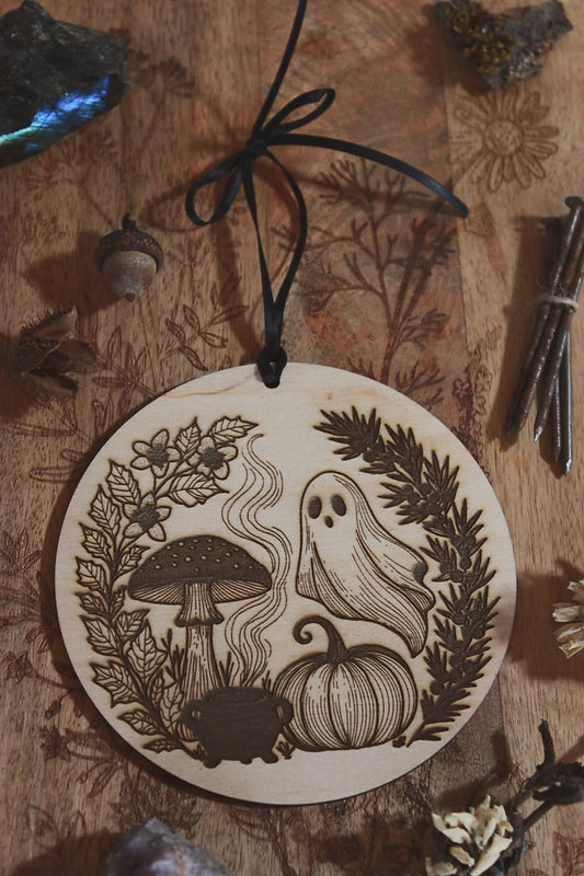 Hochkant: Rundes Holzornament mit filigranem Laserdesign: Neben Geist, Kürbis und Fliegenpilz steht ein Hexenkessel aus dem Rauch empor steigt. Eingerahmt ist das Motiv von schwarzem Bilsenkraut und Wacholder, die typisch für die Samhain-Nacht sind. Hängend an schwarzem Band, dekorativ umgeben von Naturmaterialien wie Steinen, getrockneten Blüten und Zweigen – ideal als saisonale Deko zu Samhain oder Halloween.