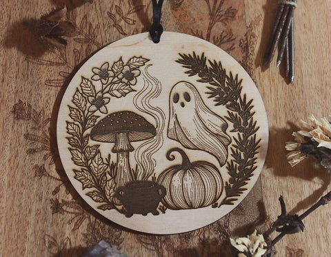 Rundes Holzornament mit filigranem Laserdesign: Neben Geist, Kürbis und Fliegenpilz steht ein Hexenkessel aus dem Rauch empor steigt. Eingerahmt ist das Motiv von schwarzem Bilsenkraut und Wacholder, die typisch für die Samhain-Nacht sind. Hängend an schwarzem Band, dekorativ umgeben von Naturmaterialien wie Steinen, getrockneten Blüten und Zweigen – ideal als saisonale Deko zu Samhain oder Halloween.