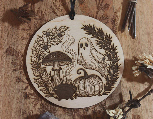 Rundes Holzornament mit filigranem Laserdesign: Neben Geist, Kürbis und Fliegenpilz steht ein Hexenkessel aus dem Rauch empor steigt. Eingerahmt ist das Motiv von schwarzem Bilsenkraut und Wacholder, die typisch für die Samhain-Nacht sind. Hängend an schwarzem Band, dekorativ umgeben von Naturmaterialien wie Steinen, getrockneten Blüten und Zweigen – ideal als saisonale Deko zu Samhain oder Halloween.