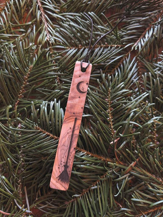 Ein Lesezeichen aus Holz in das ein Hexenbesen und ein Mond eingraviert sind.