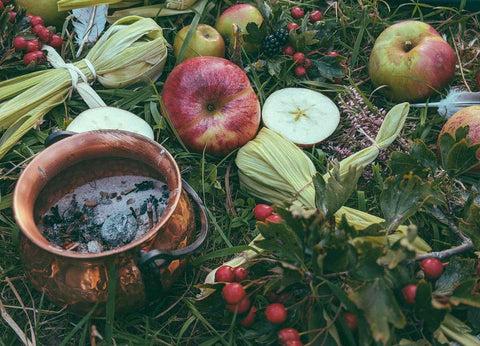 Eine Naturszene im Gras, man sieht Äpfel und Beeren neben einem Kupferkessel in dem geräuchert wird.