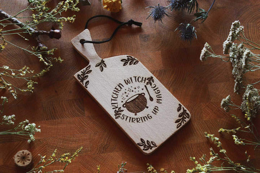 Von Kräutern umrahmt, liegt ein kleines Holzbrett auf einer Holzoberfläche. Auf dem Schneidebrettchen ist ein Hexenkessel eingebrannt. Drumherum steht geschrieben: Kitchen Witch Stirring up magic
