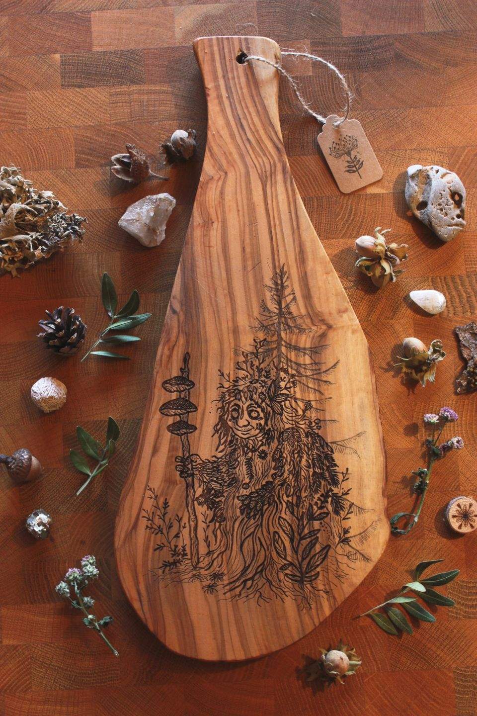 Olivenholzbrett mit eingebrannter Illustration eines Moosweibleins: eine freundliche, waldige Gestalt mit Laub, Moos und Pilzen am Stab, umgeben von Naturmaterialien wie Zapfen, Blättern und Steinen.