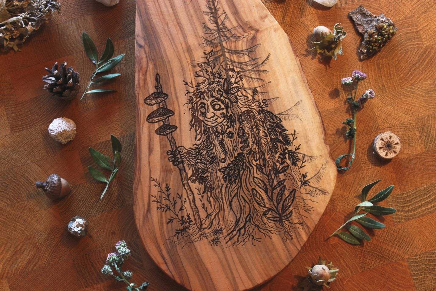 Nähere Aufnahme: Olivenholzbrett mit eingebrannter Illustration eines Moosweibleins: eine freundliche, waldige Gestalt mit Laub, Moos und Pilzen am Stab, umgeben von Naturmaterialien wie Zapfen, Blättern und Steinen.