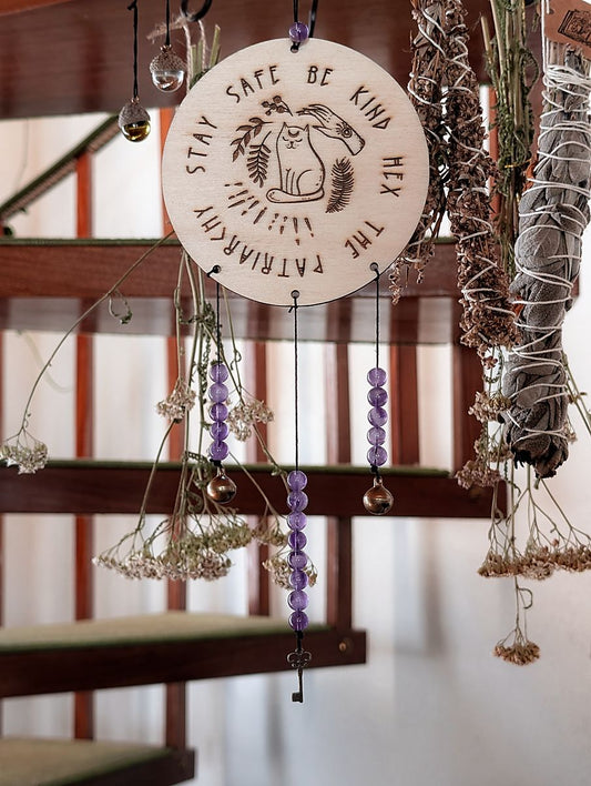 An einem Haken hängt eine Holzscheibe auf der ein Motiv eingebrannt ist. Es zeigt eine Katze, eine Hand und einige Pflanzen. Drumherum steht der Spruch "Stay safe be kind hex the patriarchy" An der Holzscheibe baumeln Glöckchen, ein Schlüssel und Amethyst Perlen.