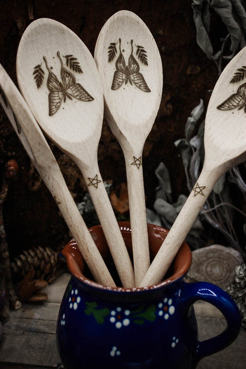 Mehrere handgeschnitzter Holzlöffel mit kunstvoller Schmetterlings- und Blattgravur stecken in einer blauen Kanne, präsentiert auf einem rustikalen Hintergrund mit getrockneten Blumen und Kiefernzapfen. 
