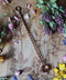 Das Bild zeigt eine rustikale, künstlerische Komposition auf einer Holzoberfläche mit filigranen floralen Gravuren. Darauf arrangiert sind verschiedene getrocknete Blumen und Kräuter wie Lavendel, Rosen und andere bunte Blüten. In der Mitte des Bildes liegt ein langes, gedrehtes Metalllöffelchen aus Kupfer mit einer kleinen, runden Schale an einem Ende. Die Kombination der Blumen, Kräuter und des Löffelchens auf der kunstvollen Holzoberfläche schafft eine harmonische, ästhetische Stimmung.