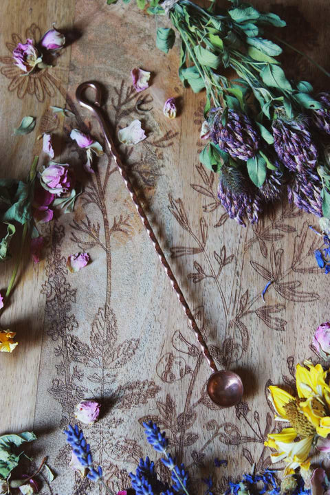 Das Bild zeigt eine rustikale, künstlerische Komposition auf einer Holzoberfläche mit filigranen floralen Gravuren. Darauf arrangiert sind verschiedene getrocknete Blumen und Kräuter wie Lavendel, Rosen und andere bunte Blüten. In der Mitte des Bildes liegt ein langes, gedrehtes Metalllöffelchen aus Kupfer mit einer kleinen, runden Schale an einem Ende. Die Kombination der Blumen, Kräuter und des Löffelchens auf der kunstvollen Holzoberfläche schafft eine harmonische, ästhetische Stimmung.