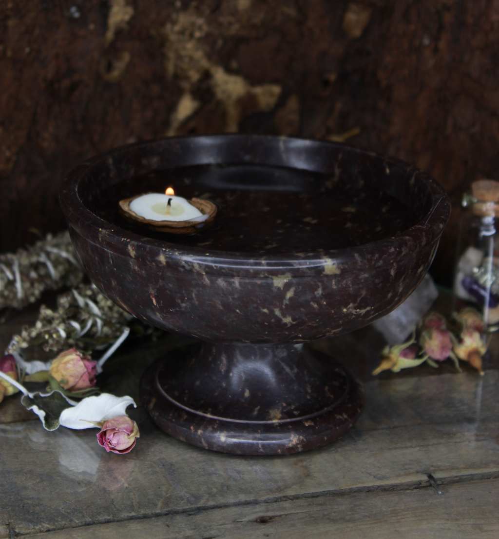 Mit Wasser gefüllte Schale aus Stein mit brennender Kerze in Walnussschale darin, umgeben von getrockneten Blumen auf rustikalem Holztisch.