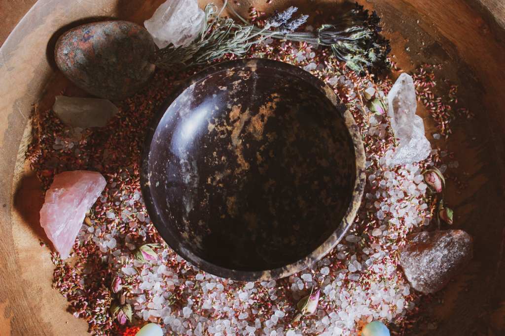 Atmosphärisches Bild einer dunklen, mystischen Steinschale, umgeben von farbenfrohen Edelsteinen und Salzen und getrockneten Blumen auf einem hölzernen Untergrund