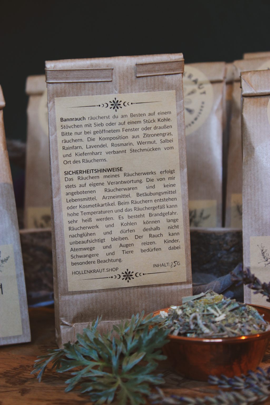 Auf einem Holztisch steht eine Kupferschale mit dem Bannrauch Räucherwerk gegen Mücken und zur Insektenabwehr darin. Drumherum liegen Kräuter wie Wermut, Salbei und Lavendel. Daneben steht ein Papierbeutel mit Sicherheitshinweisen zur Benutzung der Räuchermischung.