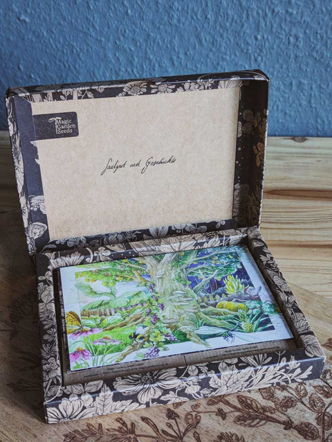Saatgut Geschenkset in einer schön illustrierten Pappverpackung mit Kräutern und Insekten. Darauf steht: Magic Garden Seeds Samenset Aromatische Kräuter für das rituelle Räuchern. Aufgeklappter Karton mit Aufdruck "Saatgut mit Geschichte". Eine Postkarte mit Baum-Motiv ist zu sehen.