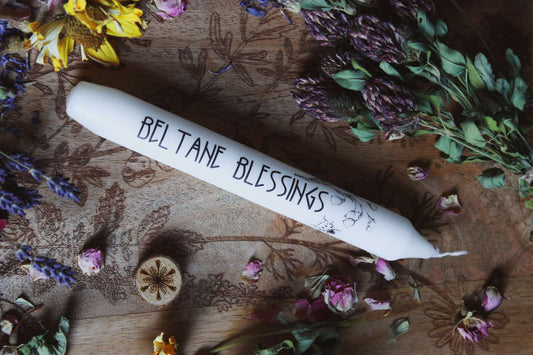 Das Bild zeigt eine weiße Kerze mit der Aufschrift „BELTANE BLESSINGS“ und dem Motiv von Maiglöckchen und einem Marienkäfer. Die Kerze liegt auf einer Holzfläche, die mit floralen Gravuren verziert ist. Rund um die Kerze sind getrocknete Blumen arrangiert, darunter gelbe, violette und pinke Blüten, sowie grüne Blätter.