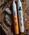 Zwei Stabkerzen liegen auf einem Holzbrett. Drumherum Steine und Halloween-Streudeko. Die obere Kerze ist gelb orange und weiß. Auf ihr steht in schwarzer Schrift: Trick or Treat
Die untere Kerze ist weiß und trägt das Motiv eines schwarzen Hexenhuts. In schwarzer Schrift steht darauf: Happy Halloween