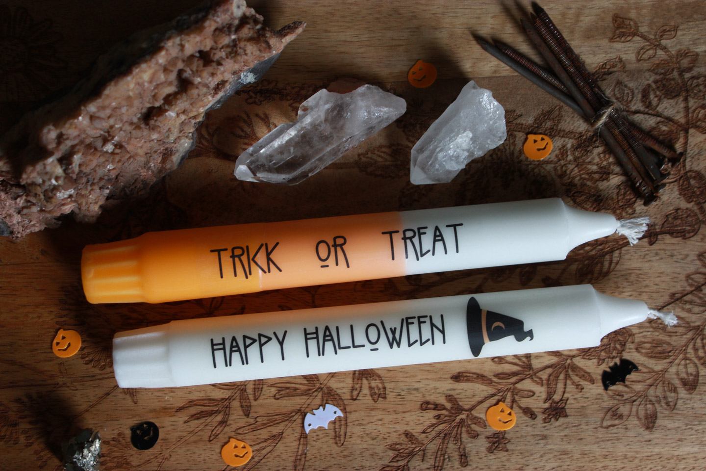 Zwei Stabkerzen liegen auf einem Holzbrett. Drumherum Steine und Halloween-Streudeko. Die obere Kerze ist gelb orange und weiß. Auf ihr steht in schwarzer Schrift: Trick or Treat
Die untere Kerze ist weiß und trägt das Motiv eines schwarzen Hexenhuts. In schwarzer Schrift steht darauf: Happy Halloween