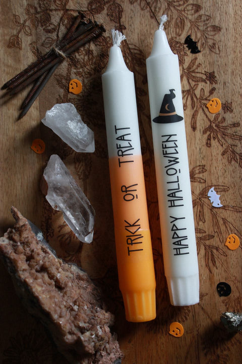 Zwei Stabkerzen liegen auf einem Holzbrett. Drumherum Steine und Halloween-Streudeko. Die obere Kerze ist gelb orange und weiß. Auf ihr steht in schwarzer Schrift: Trick or Treat
Die untere Kerze ist weiß und trägt das Motiv eines schwarzen Hexenhuts. In schwarzer Schrift steht darauf: Happy Halloween