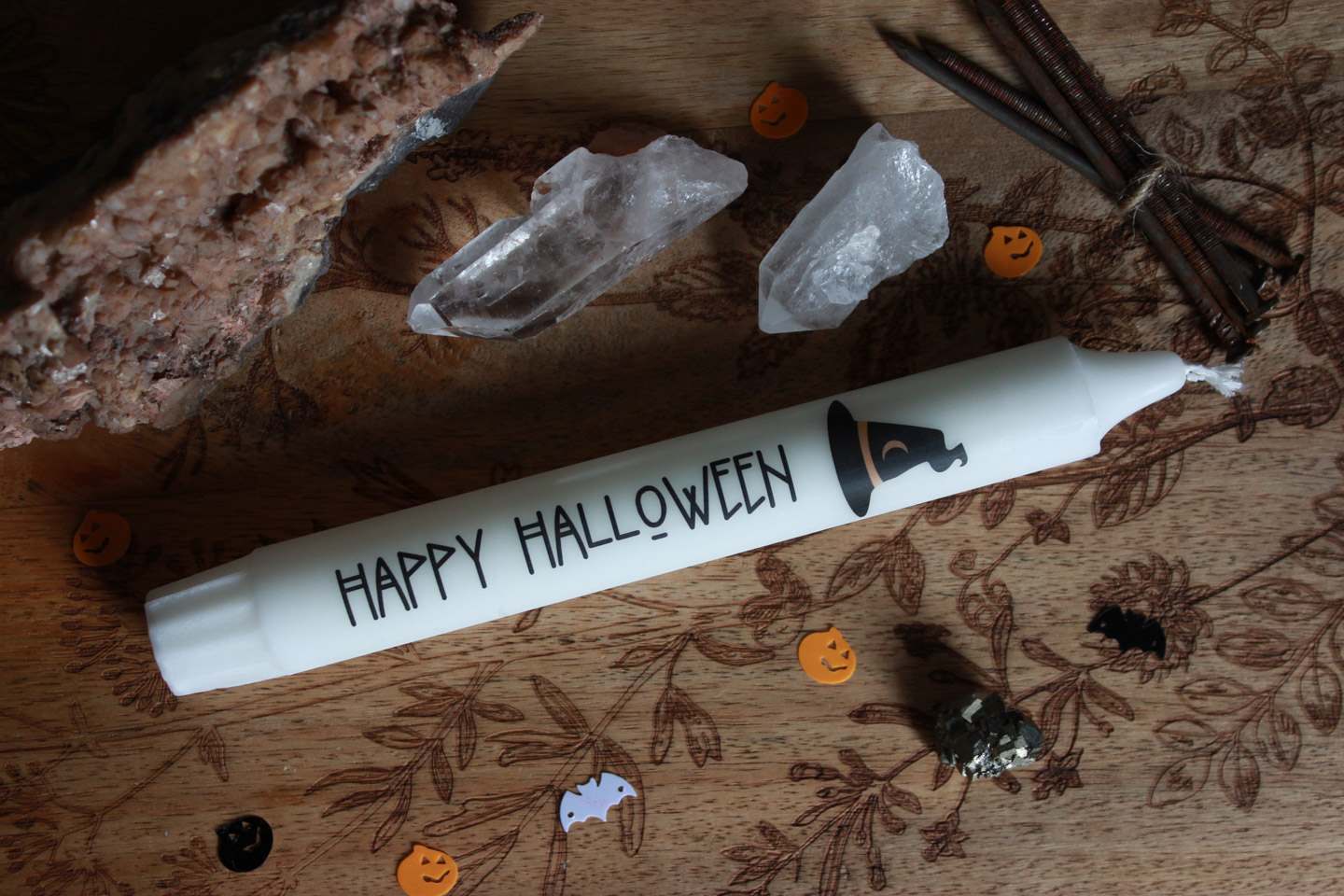 Eine Stabkerze liegt auf einem Holzbrett. Drumherum Steine und Halloween-Streudeko.
Die Kerze ist weiß und trägt das Motiv eines schwarzen Hexenhuts. In schwarzer Schrift steht darauf: Happy Halloween