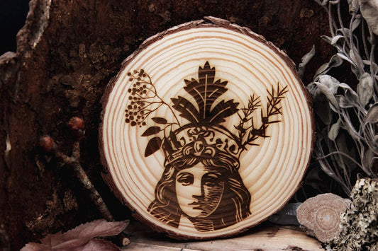 Eine Baumscheibe in der das Portait einer Frau eingebrannt ist. Eine ihrer Gesichtshälften ist dunkler gezeichnet als die andere. Auf dem Kopf trägt sie eine Krone aus Holunder, Beifuß und Wacholder.