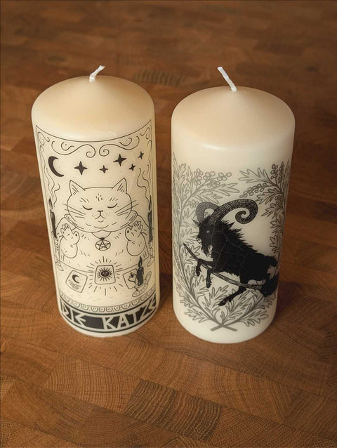 Zwei Stumpenkerzen stehen auf einem Holz-Tisch. Die linke Kerze zeigt ein Motiv mit einer Katze, die Tarotkarten legt und das rechte einen Ziegenbock der auf einem Besen reitet.