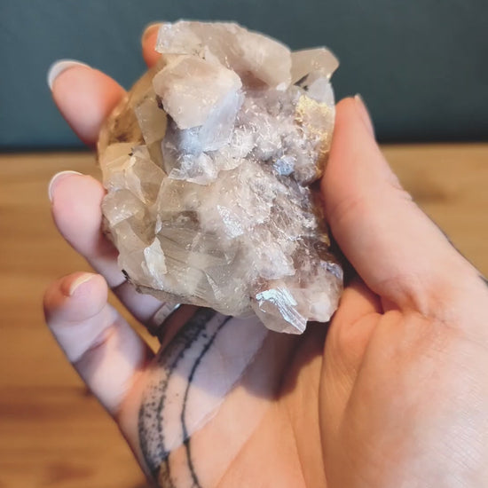 Das Video zeigt ein Stück Calcit, das in einer Hand gehalten wird. Der Calcit hat eine weiße bis leicht rosa Farbe und besteht aus mehreren kristallinen Strukturen, die in verschiedene Richtungen zeigen. Die Oberfläche des Calcits ist teilweise durchsichtig und weist einige Bruchstellen auf, die die kristalline Struktur des Minerals sichtbar machen.