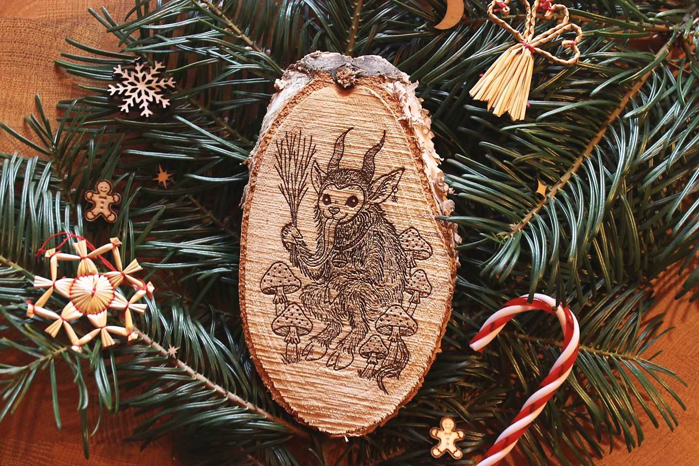 Gebettet auf Tannenzweigen und weihnachtlicher Dekoration liegt in der Mitte eine ovale Birkenholzscheibe mit eingebranntem Bild. Das Motiv zeigt einen Krampus, der zwischen Fliegenpilzen sitzt. Er hat einen Pentagramm-Ohrring am Ohr, eine große Glocke um den Hals und hält eine Rute aus Zweigen in der Klaue.