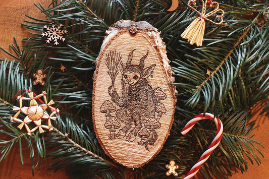Gebettet auf Tannenzweigen und weihnachtlicher Dekoration liegt in der Mitte eine ovale Birkenholzscheibe mit eingebranntem Bild. Das Motiv zeigt einen Krampus, der zwischen Fliegenpilzen sitzt. Er hat einen Pentagramm-Ohrring am Ohr, eine große Glocke um den Hals und hält eine Rute aus Zweigen in der Klaue.