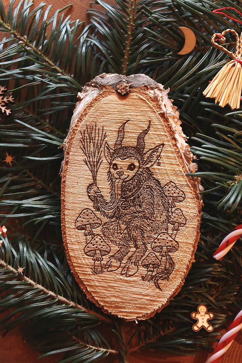 Hochkant: Gebettet auf Tannenzweigen und weihnachtlicher Dekoration liegt in der Mitte eine ovale Birkenholzscheibe mit eingebranntem Bild. Das Motiv zeigt einen Krampus, der zwischen Fliegenpilzen sitzt. Er hat einen Pentagramm-Ohrring am Ohr, eine große Glocke um den Hals und hält eine Rute aus Zweigen in der Klaue.