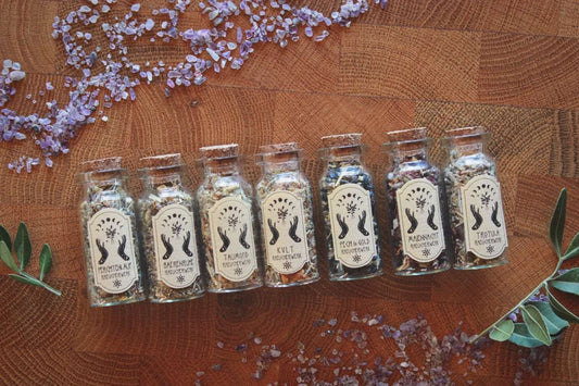 Sieben Glasflaschen mit Korken, gefüllt mit getrockneten Kräutern, liegen auf einem rustikalen Holztisch, umgeben von kleinen Amethyst-Steinen. Auf den Etiketten steht von links nach rechts: Perchtenlauf, Bärenruhe, Taumond, KVLT, Pech & Gold, Maiennacht und Trotula.