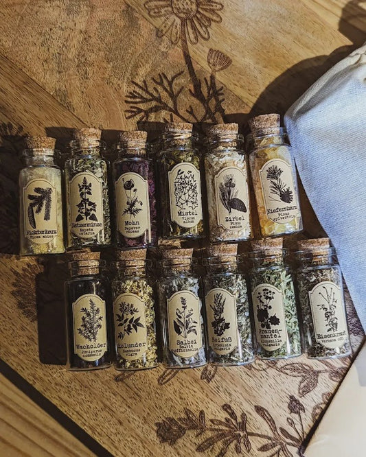 Zwölf kleine Glasfläschchen mit Korkverschluss, liebevoll arrangiert auf einem gravierten Holzbrett. Jedes Fläschchen enthält getrocknete Kräuter oder Harze und trägt ein vintage-inspiriertes Etikett mit botanischer Illustration und deutschem Pflanzennamen – darunter Katzenminze, Beifuß und Baldrian. Die Szene strahlt eine ruhige, naturverbundene Atmosphäre aus und erinnert an eine sorgfältig kuratierte Kräutersammlung.