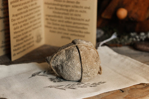 Das Bild zeigt eine in zwei Hälften geschnittene Eichenberger Kugel, die mit einer Schnur zusammen gebunden ist. Unter dem Stein liegt ein Jutebeutel. Dahinter steht aufgeklappt ein Zine. Der Text darauf ist unscharf und zu weit im Hintergrund.