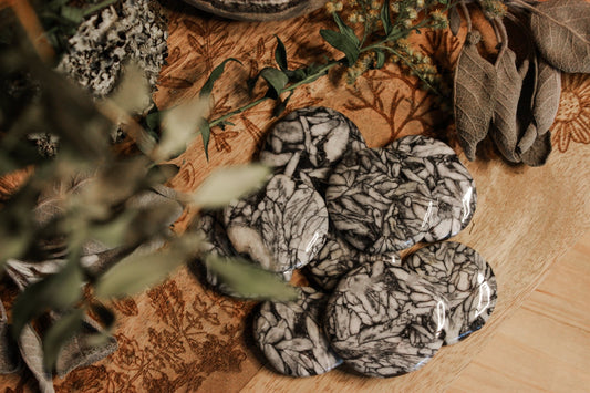 Eisblumen-Magnesit Scheibenstein aus Österreich - Faire regionale Steine Marmorierte Steine mit komplexen schwarz-weißen Mustern, platziert auf einem hölzernen Untergrund mit floralen Gravuren, umgeben von Blättern und einer Pflanze